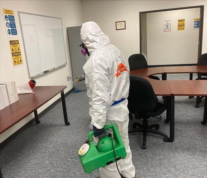 technician wearing a full personal protective suit carrying a jug of disinfectant spray